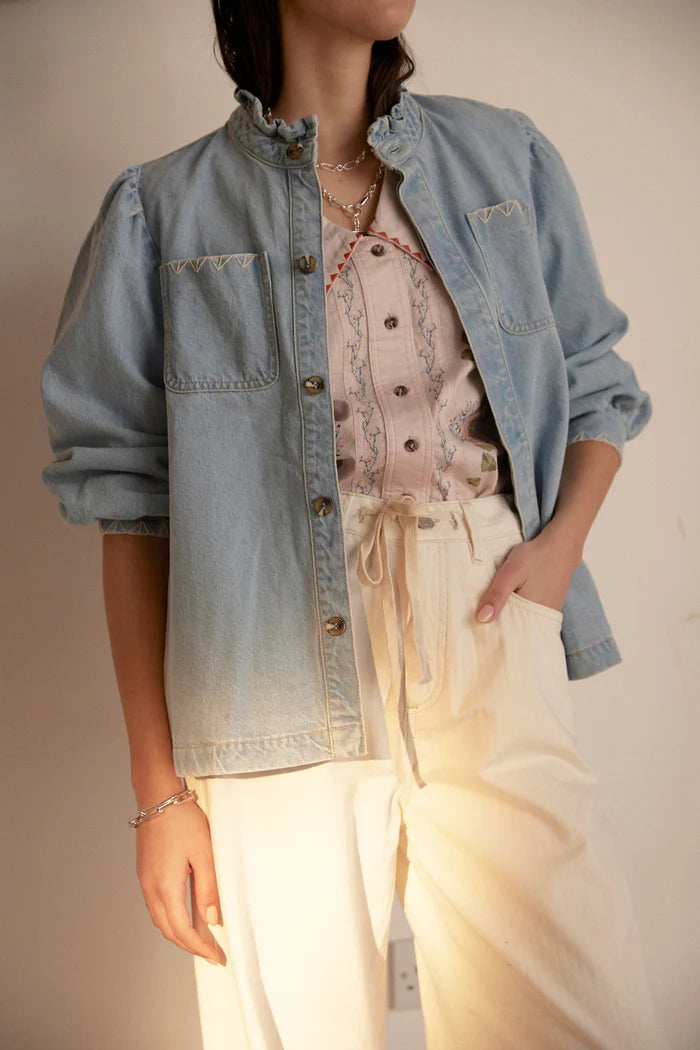 A person wearing the Seventy & Mochi Dana Denim Jacket in Sunbleached over a patterned shirt with a ruffled collar and cream drawstring pants, standing by a plain wall.