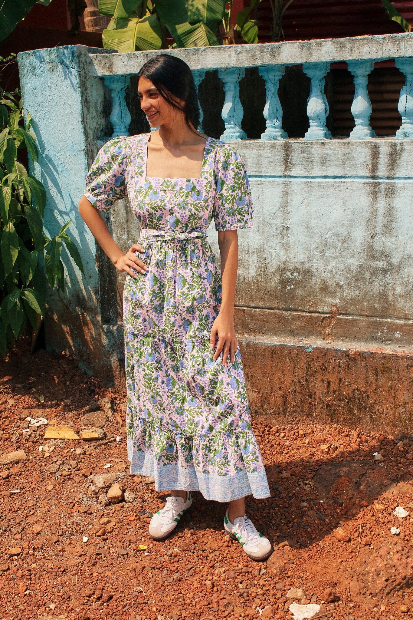 A woman stands outdoors wearing the Divya Dress - Cornflower Blue from SZ Blockprints and white sneakers. She has dark hair tied back and is posed with one hand on her hip in front of a blue wall.