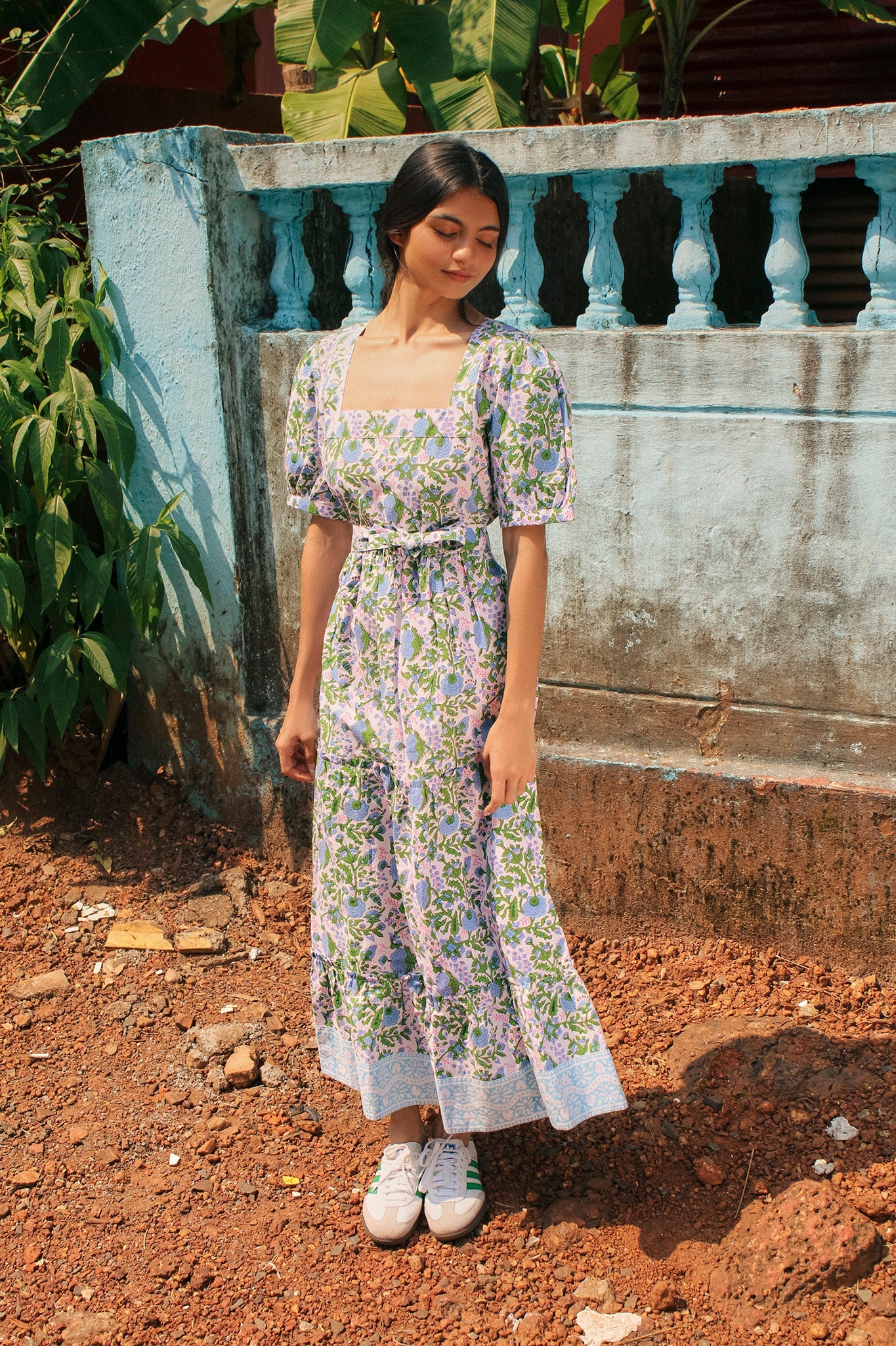 A woman in a Divya Dress - Cornflower Blue by SZ Blockprints and white sneakers stands in front of a weathered blue wall and greenery. She has a contemplative expression and is looking downwards.