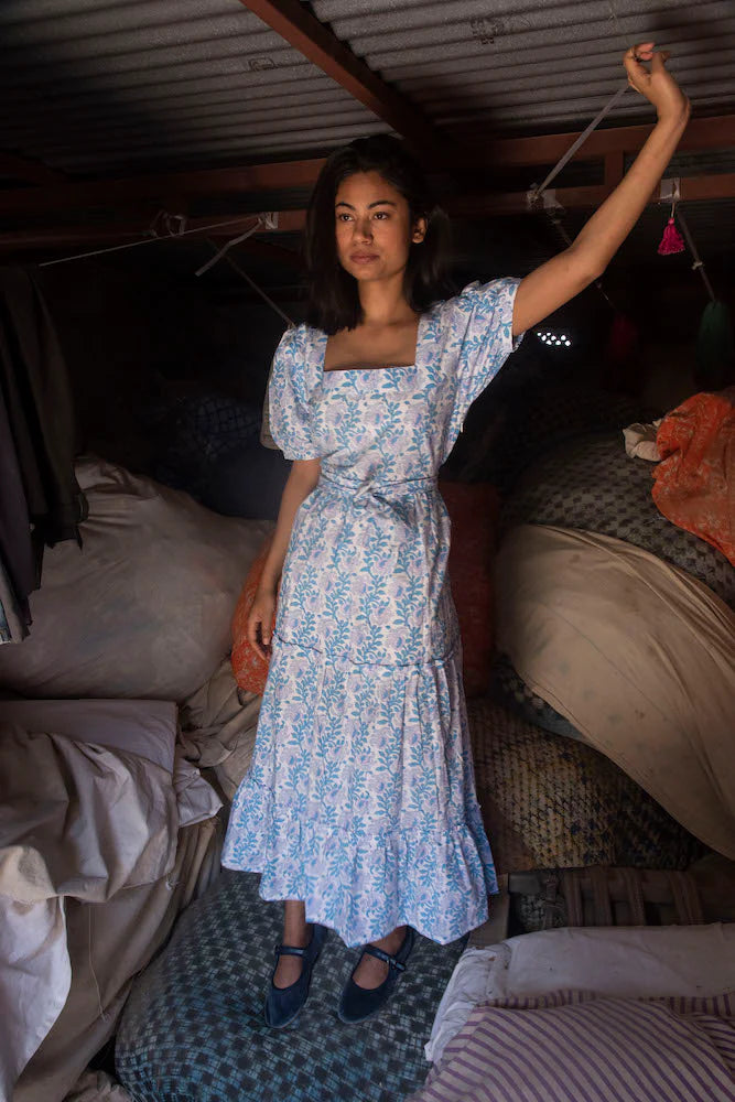 A woman in a Divya Dress - Cornflower Blue from SZ Blockprints with a square neckline stands under a low ceiling, holding onto a support, surrounded by bedding and clothes.