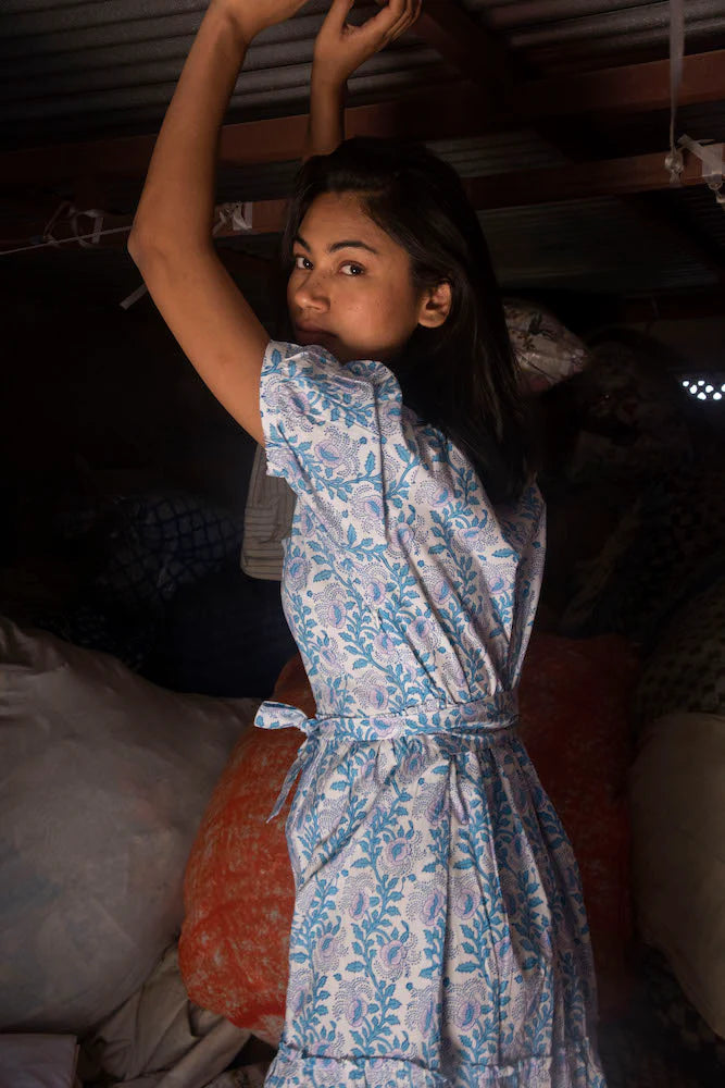 Young woman in a SZ Blockprints Divya Dress - Cornflower Blue with a square neckline, raising her arm and looking at the camera, standing in a dimly lit storage room filled with bags.