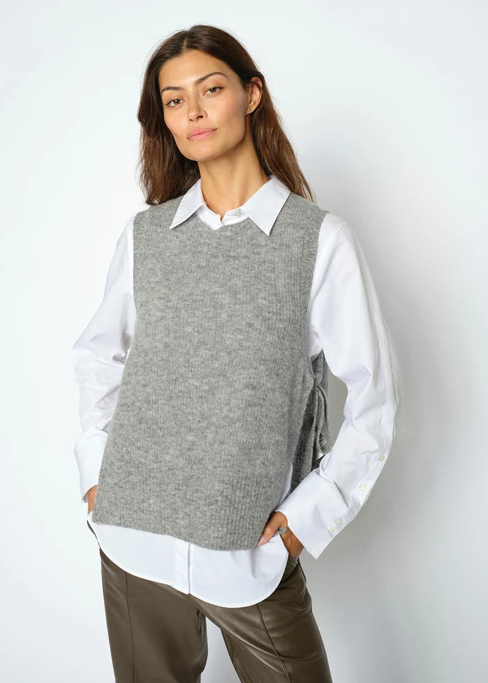 A woman stands against a plain white background wearing the MOS MOSH Thora Tie Knit Vest in Grey Melange—a soft alpaca wool blend vest with adjustable tie fastenings—layered over a white collared shirt and paired with brown pants.