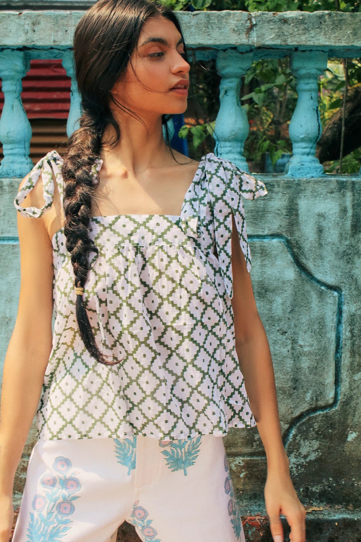A woman with a long braid stands in front of a weathered blue wall, wearing the Emma Top - Oasis Green by SZ Blockprints and light-colored pants adorned with floral designs.