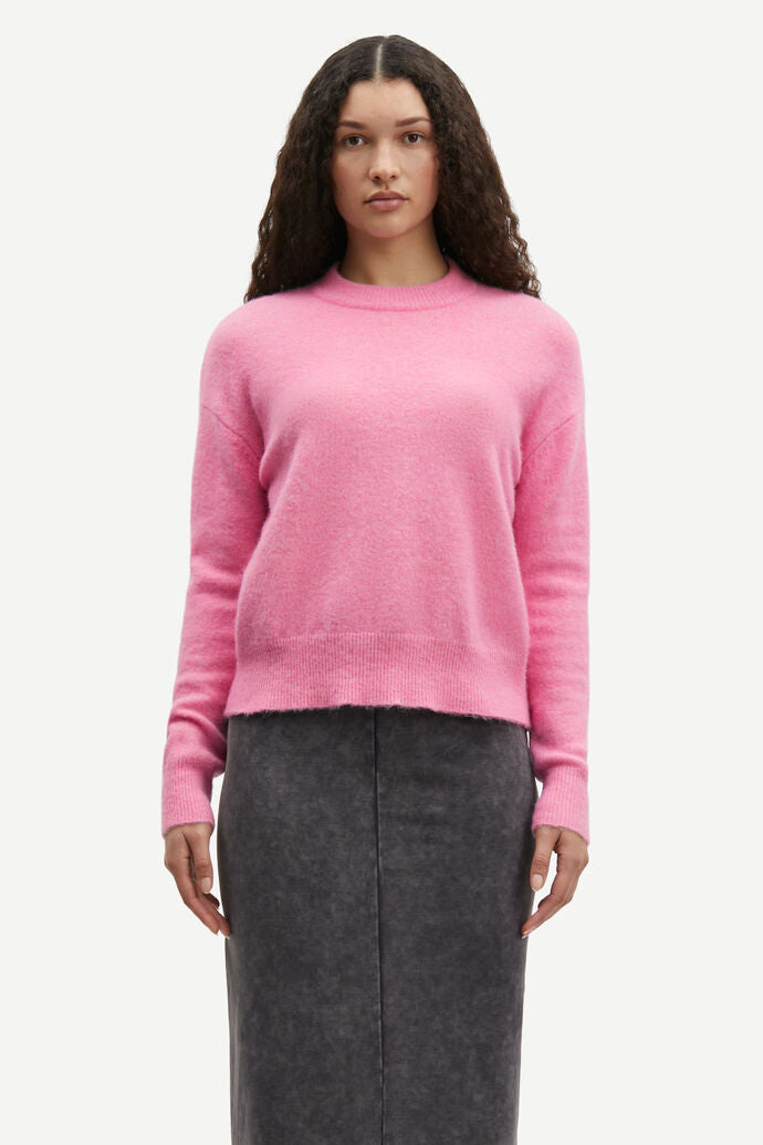 A woman with long, curly hair is wearing an Orchid Smoke Anour Sweater by Samsøe Samsøe, crafted from an elastic blend with wool and alpaca wool, paired with a dark gray skirt. She stands against a plain white background.