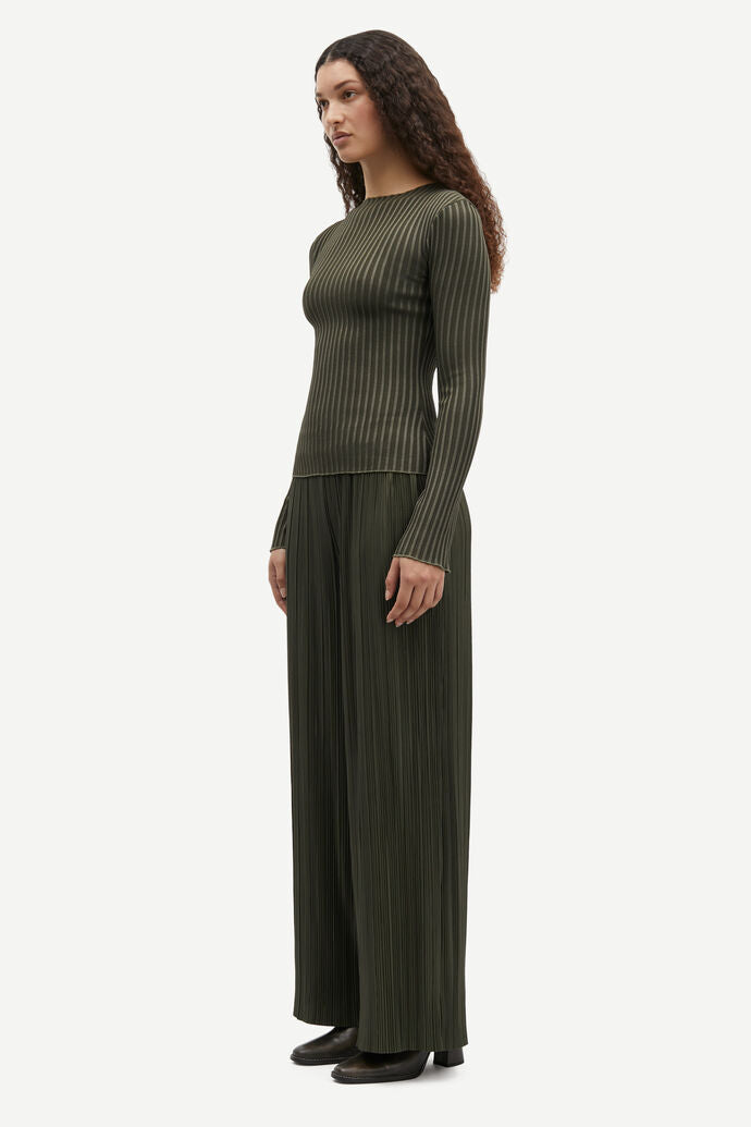 A woman with long, curly hair stands in a three-quarter view, wearing a green ribbed sweater and matching Samsøe Samsøe Uma Trousers - Forest Night made from recycled polyester, along with black shoes.