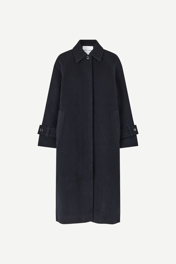 The Alma Coat - Salute by Samsøe Samsøe is a long, dark navy overcoat crafted from recycled wool. It features a collar, front buttons, and two side pockets. Additionally, the coat includes buttoned straps on the sleeves, making it ideal for winter.