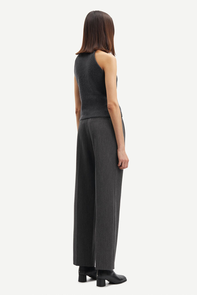 Person wears long Samsoe Samsoe trousers in a dark grey colour with a high neck tank top in the same shade. Pleated front with pockets. Side view.