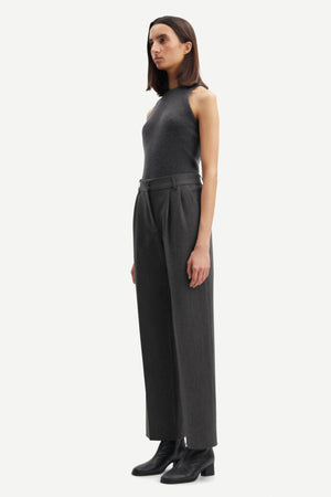 Person wears long Samsoe Samsoe trousers in a dark grey colour with a high neck tank top in the same shade. Pleated front with pockets. Side view. 
