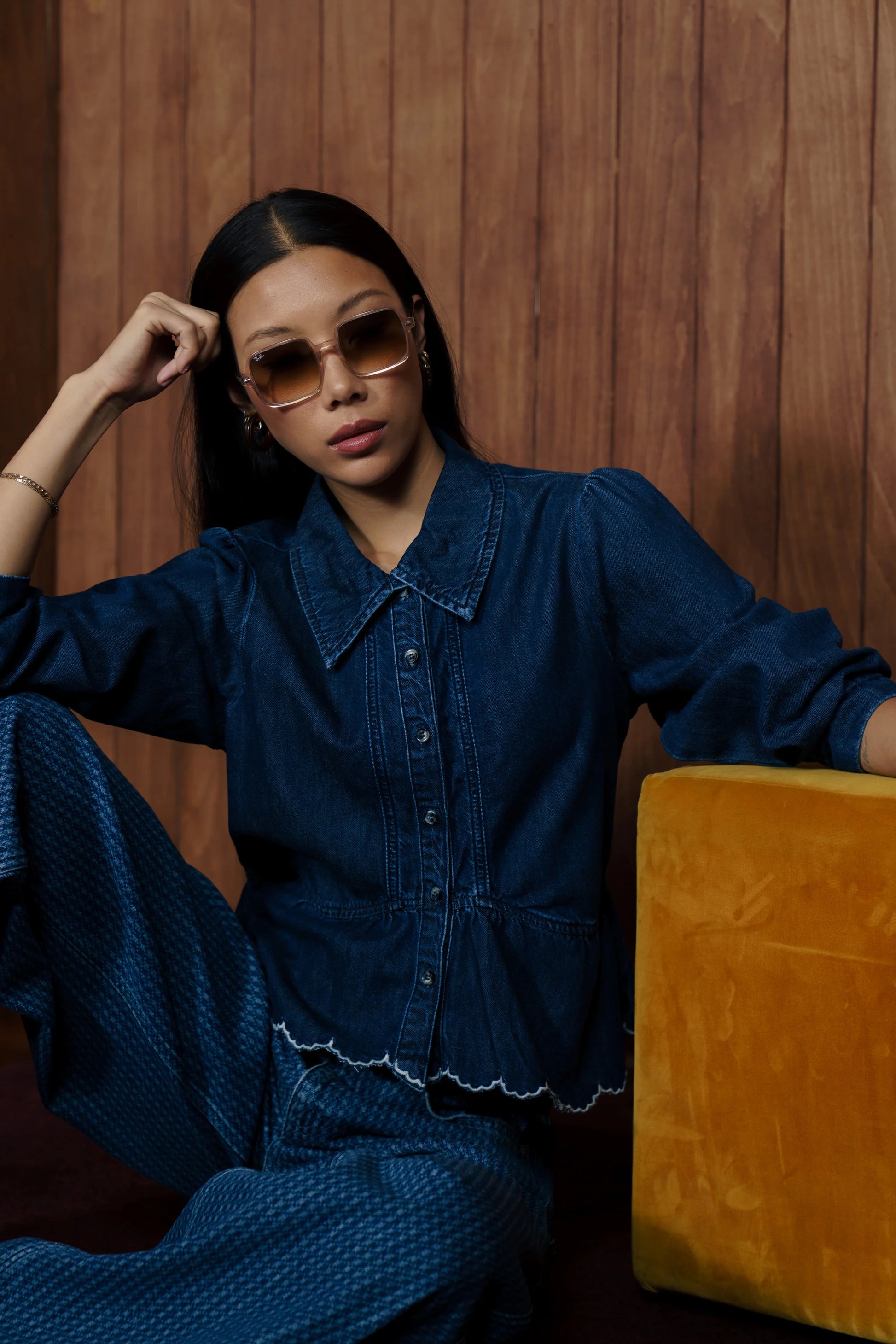 A woman wearing a dark blue denim button-up shirt and matching wide-leg pants, seated next to a mustard yellow velvet stool against a wood-paneled wall.