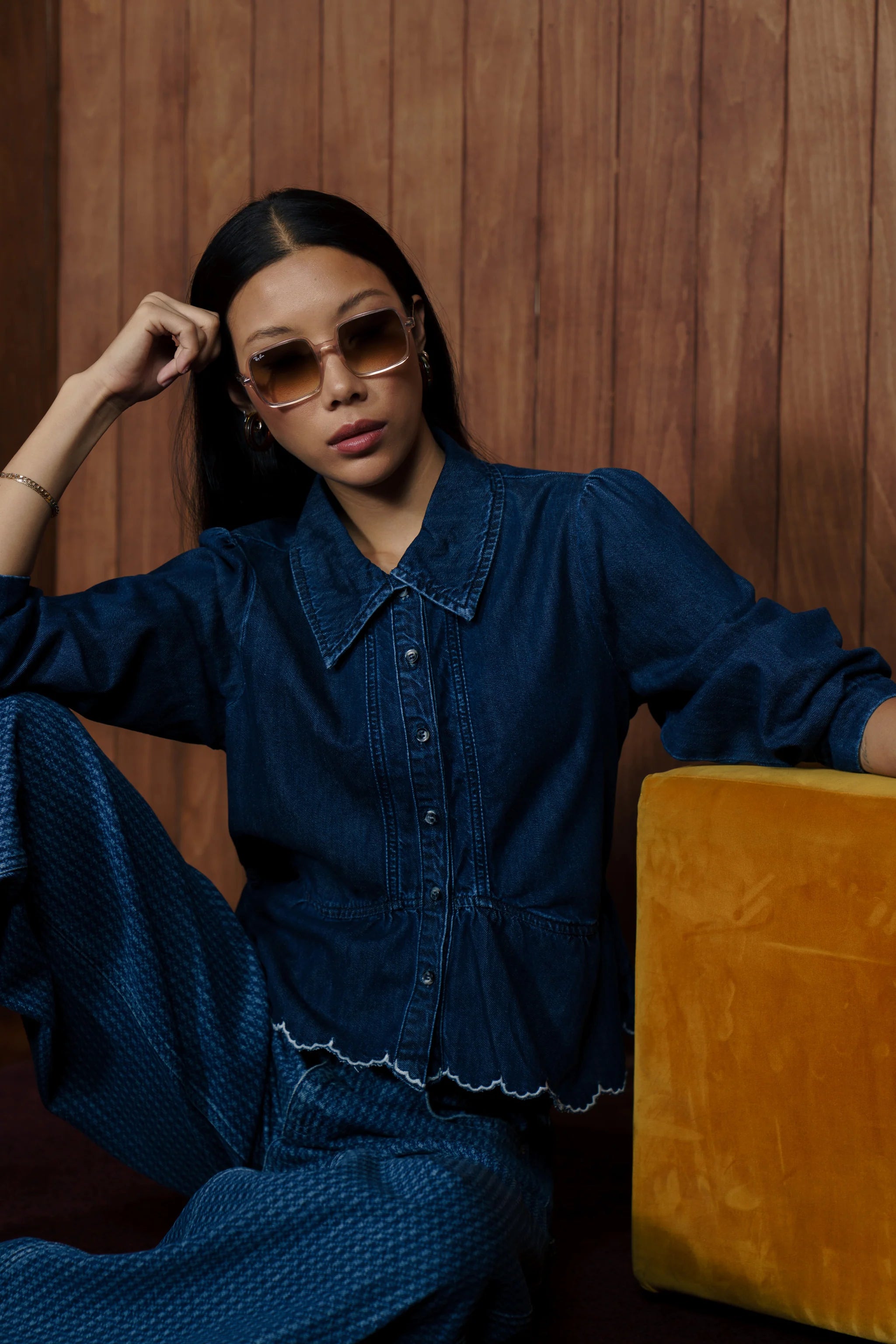 A woman wearing a dark blue denim button-up shirt and matching wide-leg pants, seated next to a mustard yellow velvet stool against a wood-paneled wall.