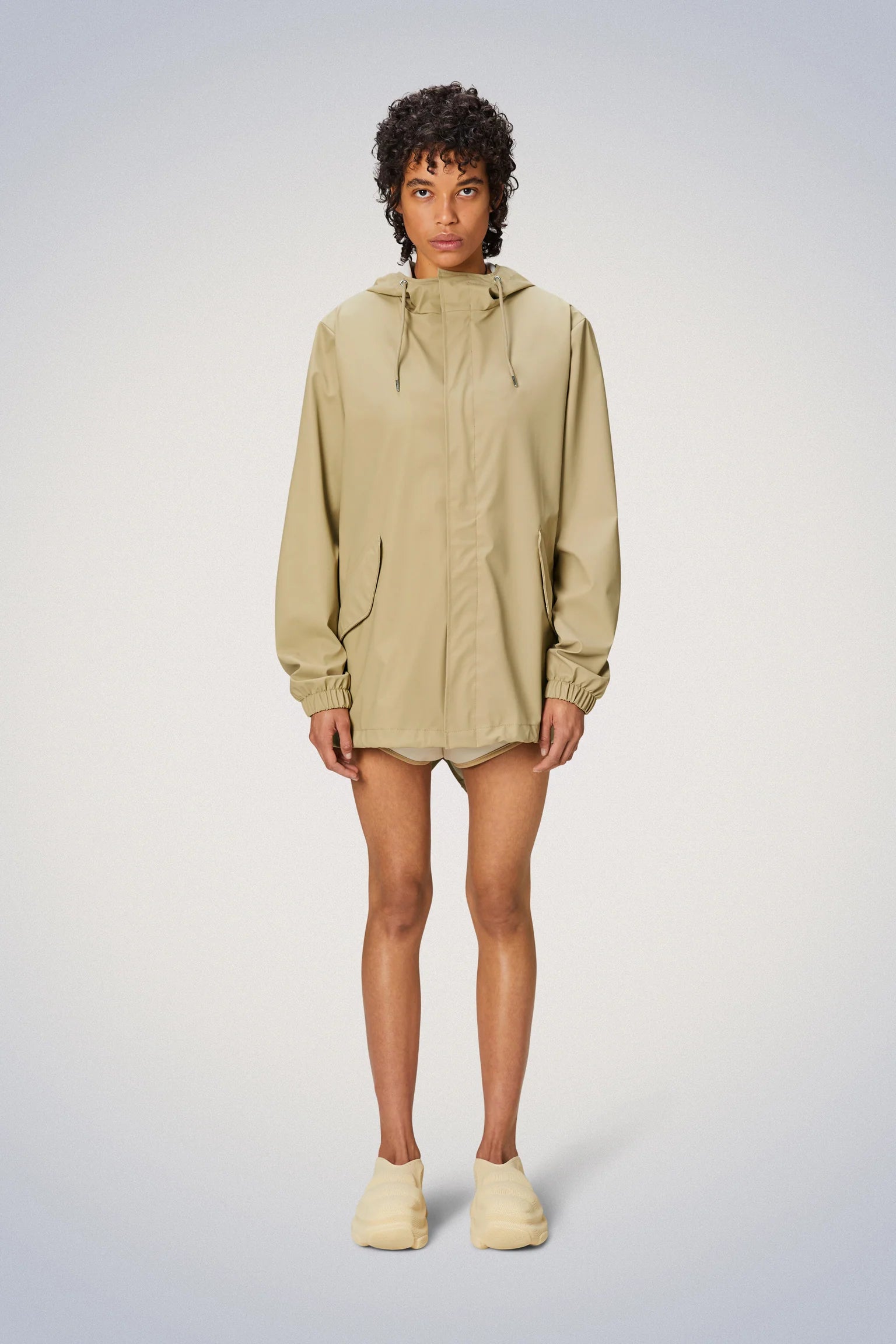 A woman wearing a Rains drawstring fishtail hem rain jacket and shorts.