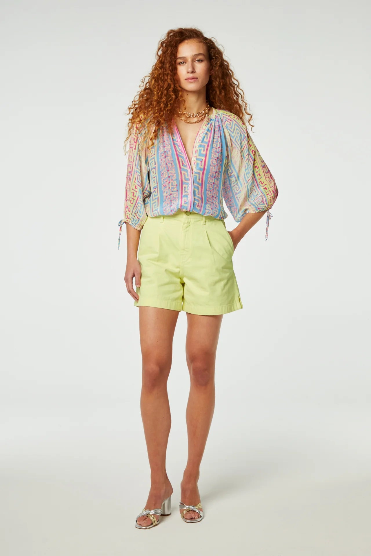 A woman stands posing in a bright yellow wash patterned blouse and lime green Fabienne Chapot Limoncello shorts, paired with silver sandals, against a white background.