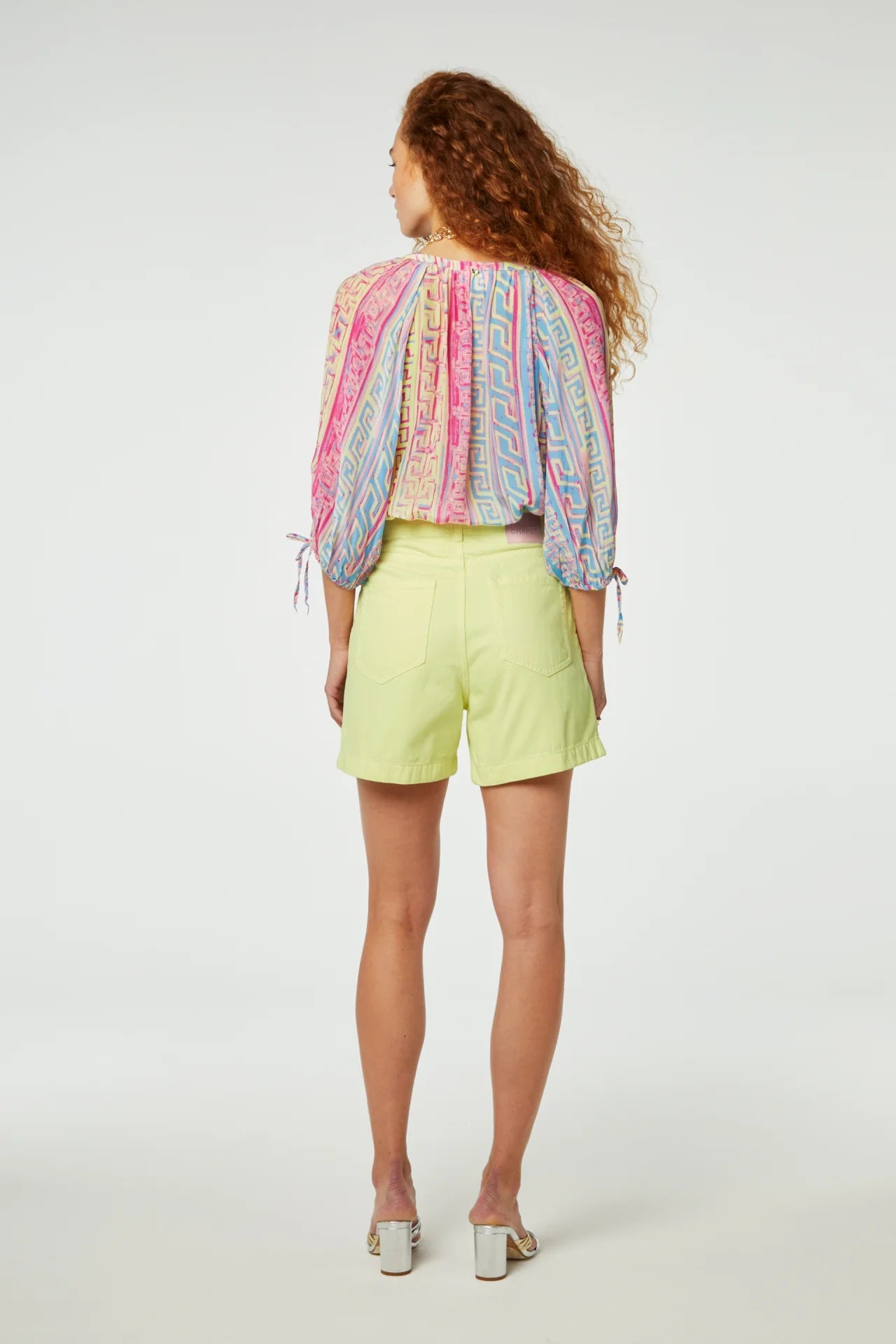 A woman stands with her back to the camera, wearing a colorful patterned blouse and Limoncello Foster shorts by Fabienne Chapot in a bright yellow wash, paired with silver heels.