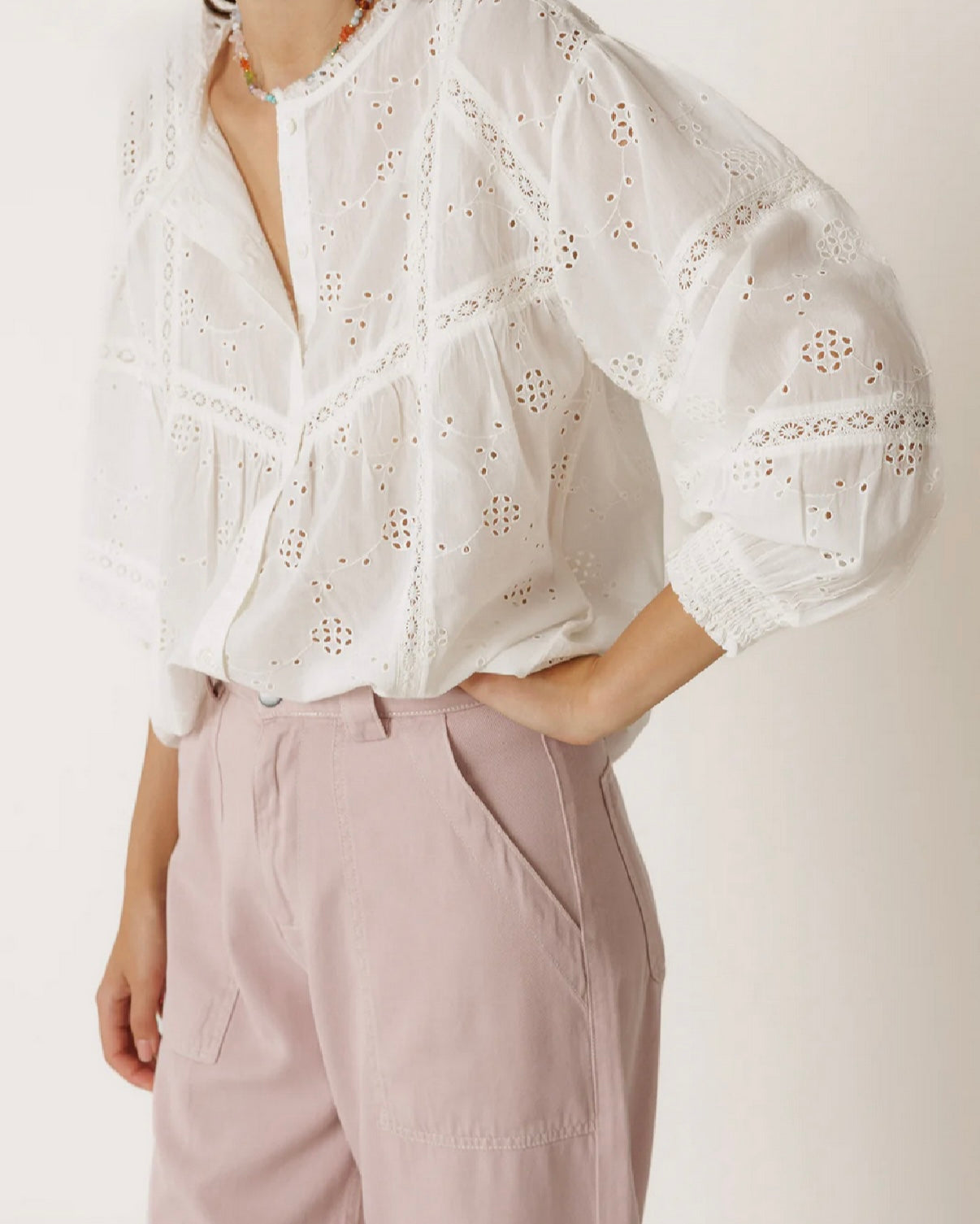 A person wears the Indi & Cold Shirt with Schiffly Embroidery in white, featuring bishop sleeves, paired with light pink high-waisted pants, standing with one hand on their hip.