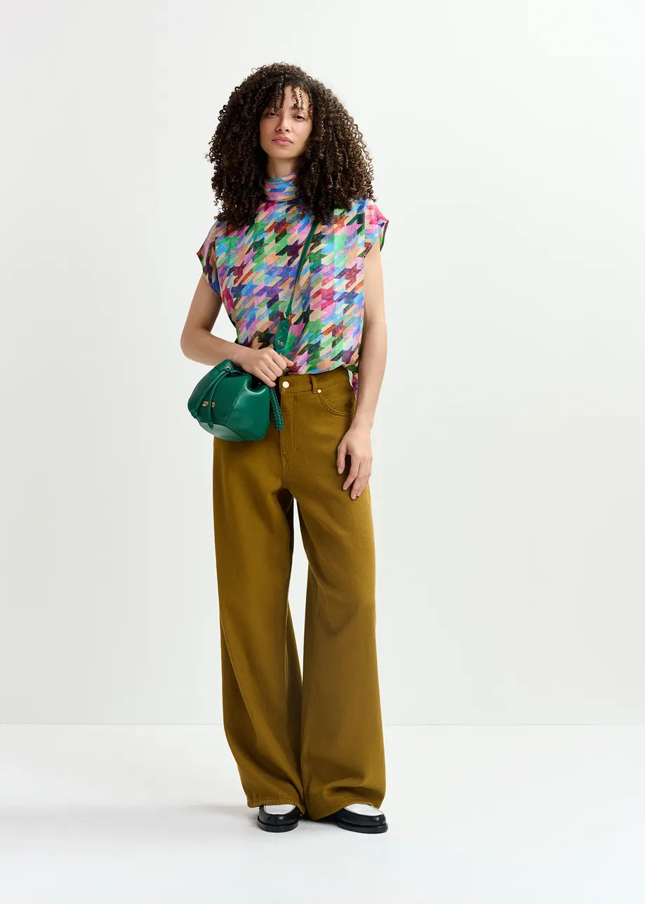 A person stands against a plain background, wearing a multicolored top, brown wide-leg pants, black shoes, and the Gabi Bag in dark green with a macramé-woven strap from Essentiel Antwerp.