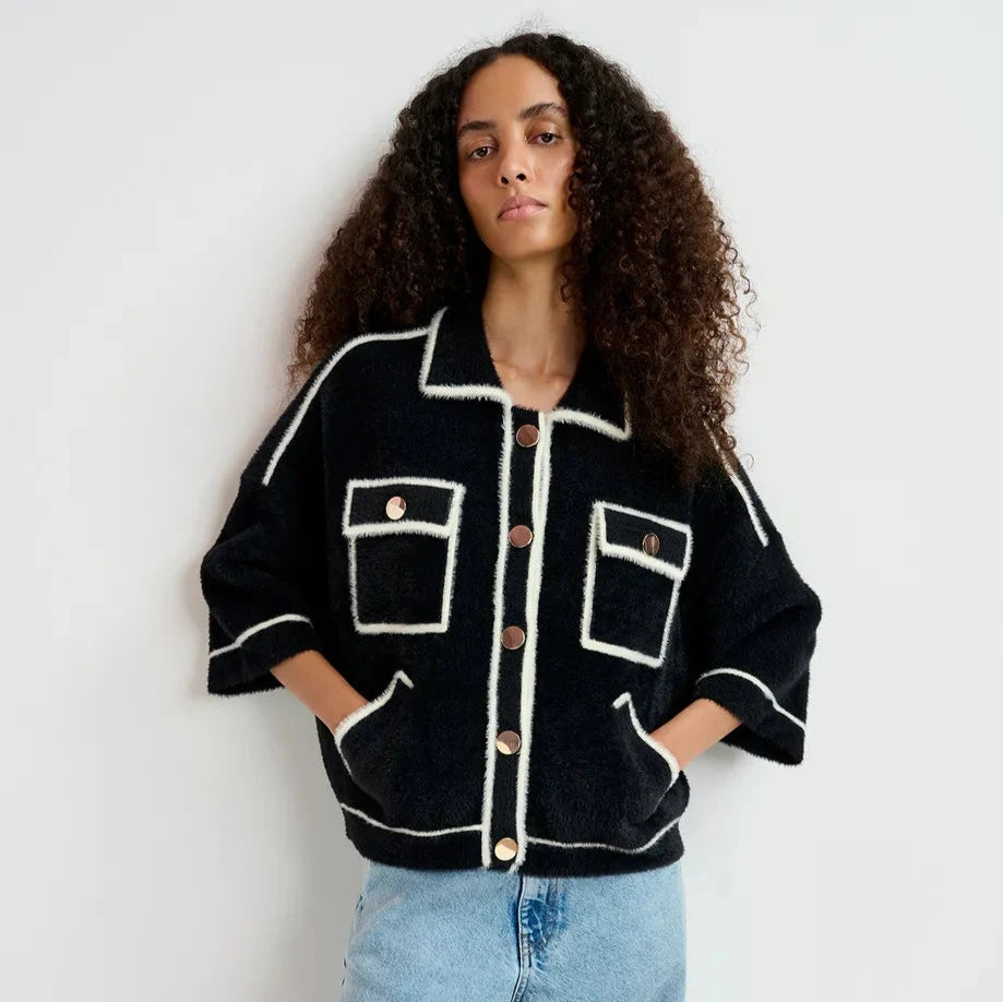 A person stands against a light-colored wall wearing the oversized 'Galena Cardigan - Black & White' by Essentiel Antwerp, featuring gold-tone buttons and pockets. They have paired it with light blue jeans, hands casually tucked in pockets.