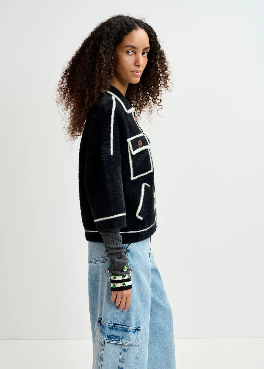 A woman with curly hair is wearing the Galena Cardigan in black and white from Essentiel Antwerp, paired with blue wide-leg pants. She also has on bracelets and a light gray top underneath. The oversized fit of her outfit complements her style as she stands sideways in a plain, light-colored room.