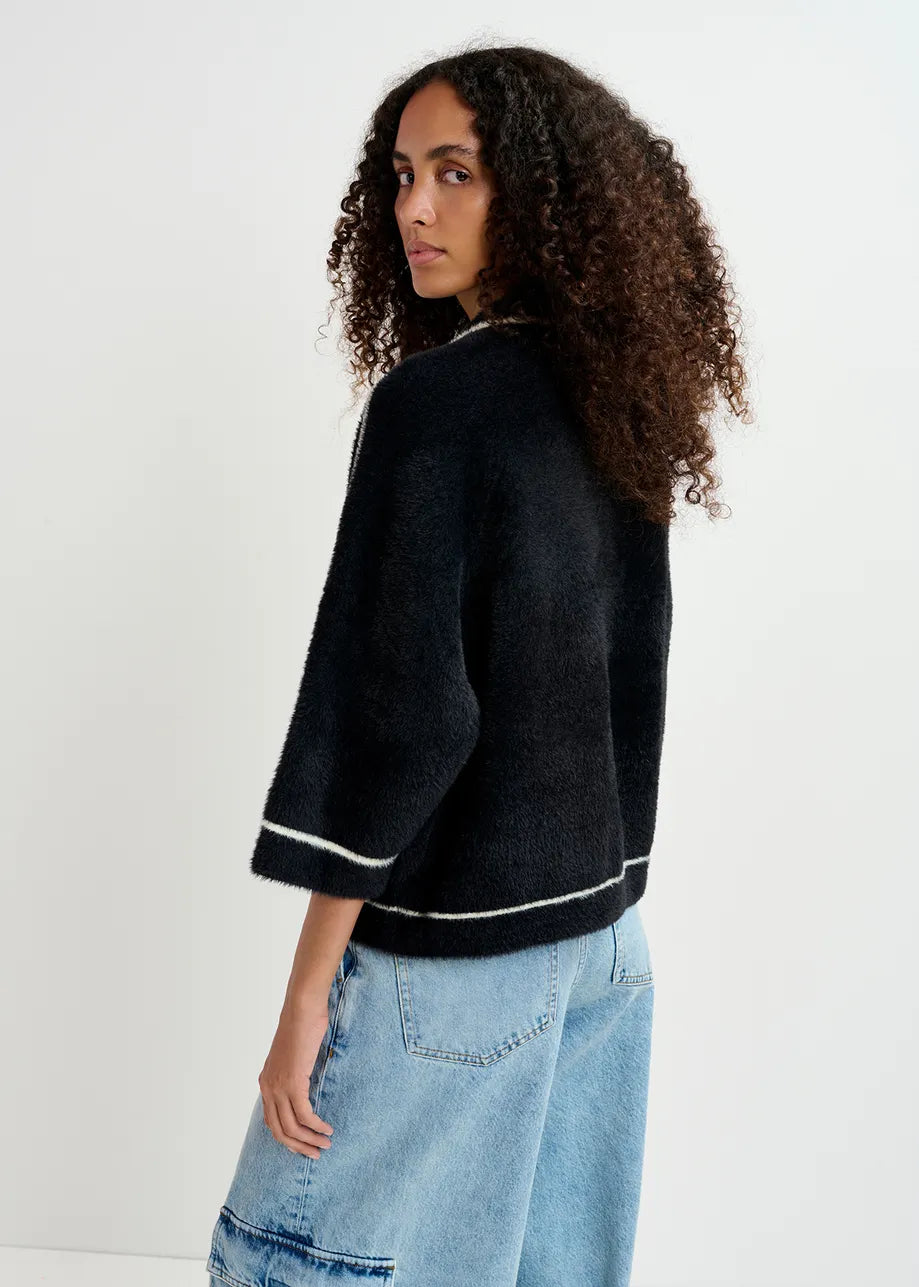 An individual with curly hair wearing the Galena Cardigan in black and white from Essentiel Antwerp, along with blue jeans, is standing and looking over their shoulder.