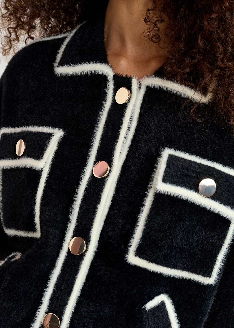 Close-up of the Essentiel Antwerp Galena Cardigan - Black & White with white trim and large gold-tone buttons. The oversized silhouette features two chest pockets with button closures. The curly hair of the wearer is partially visible.