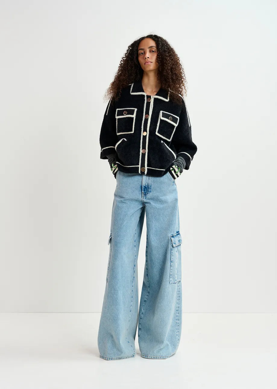 A person with curly hair stands against a plain background, wearing an oversized silhouette black and white Galena Cardigan by Essentiel Antwerp, featuring gold-tone buttons and large pockets over light blue wide-leg jeans with cargo pockets.