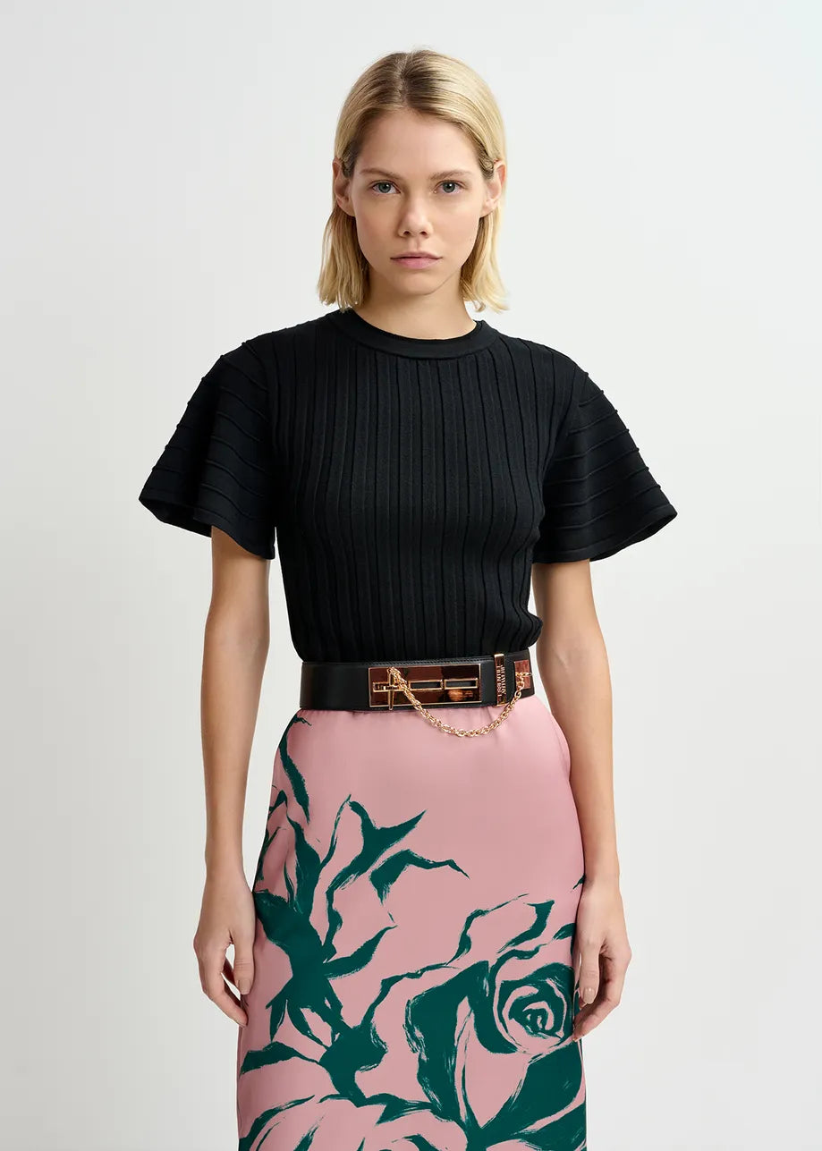 A woman with short blonde hair is wearing a black ribbed short-sleeve top and a pink skirt with a large green floral print, cinched at the waist with the Essentiel Antwerp Gamme Belt in Black, featuring a gold-tone buckle.