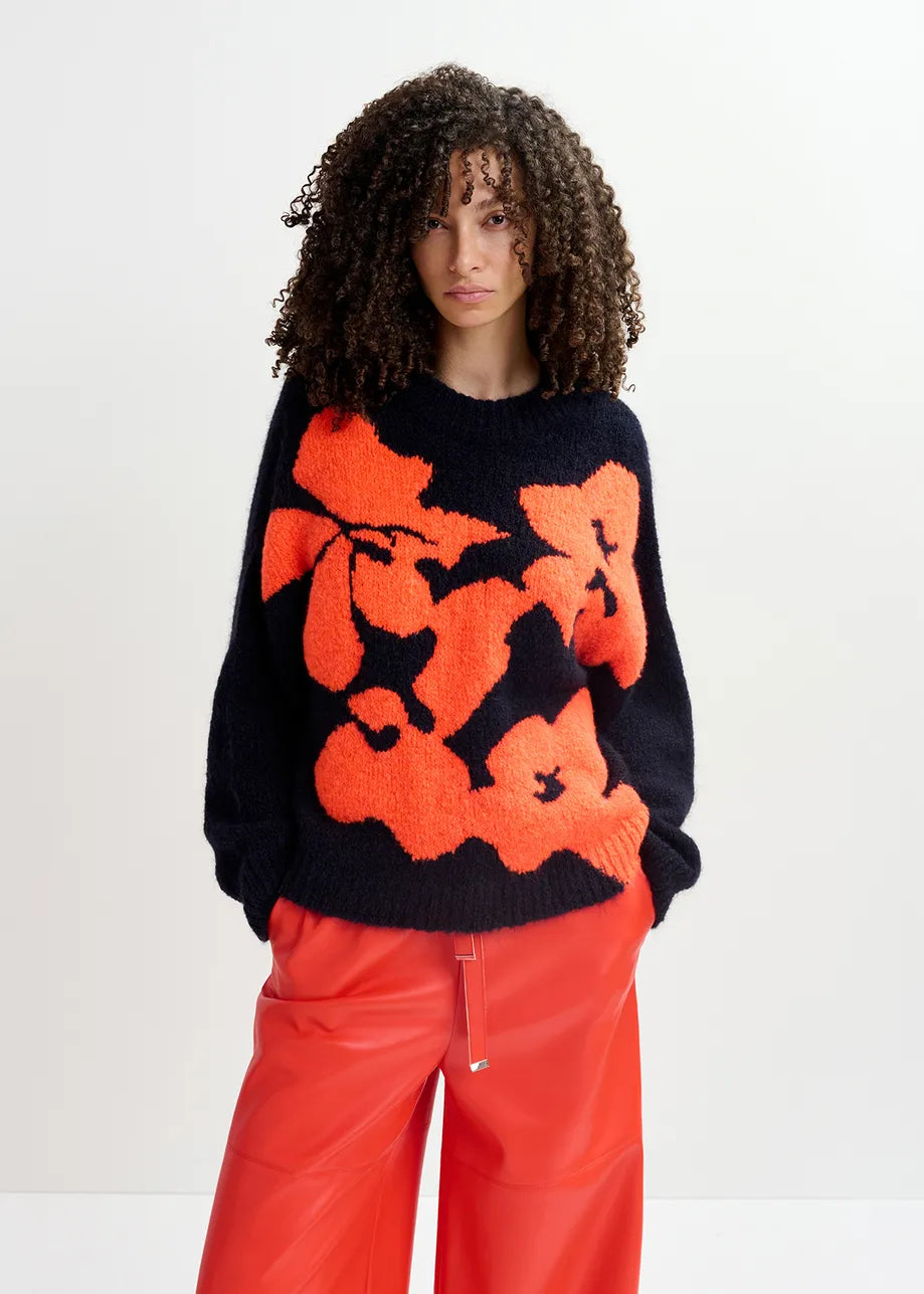 A person with curly hair is wearing an oversized Garcon Sweater in navy and red by Essentiel Antwerp, plus bright red pants.