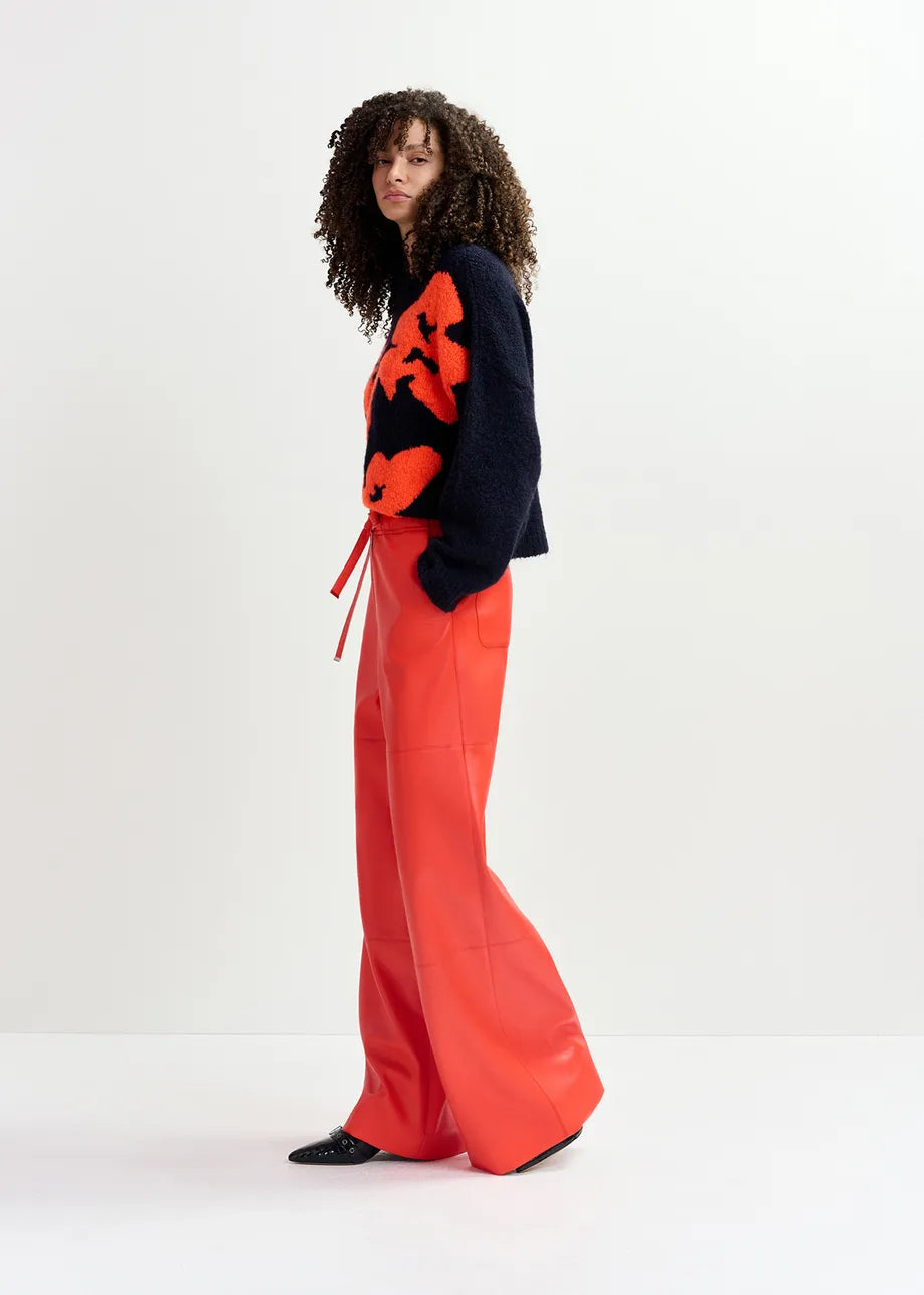 A person stands against a plain backdrop, wearing an oversized Garcon Sweater – Navy and Red by Essentiel Antwerp, featuring orange patterns resembling a floral design, paired with bright red wide-leg pants. They have curly hair and are looking towards the camera.