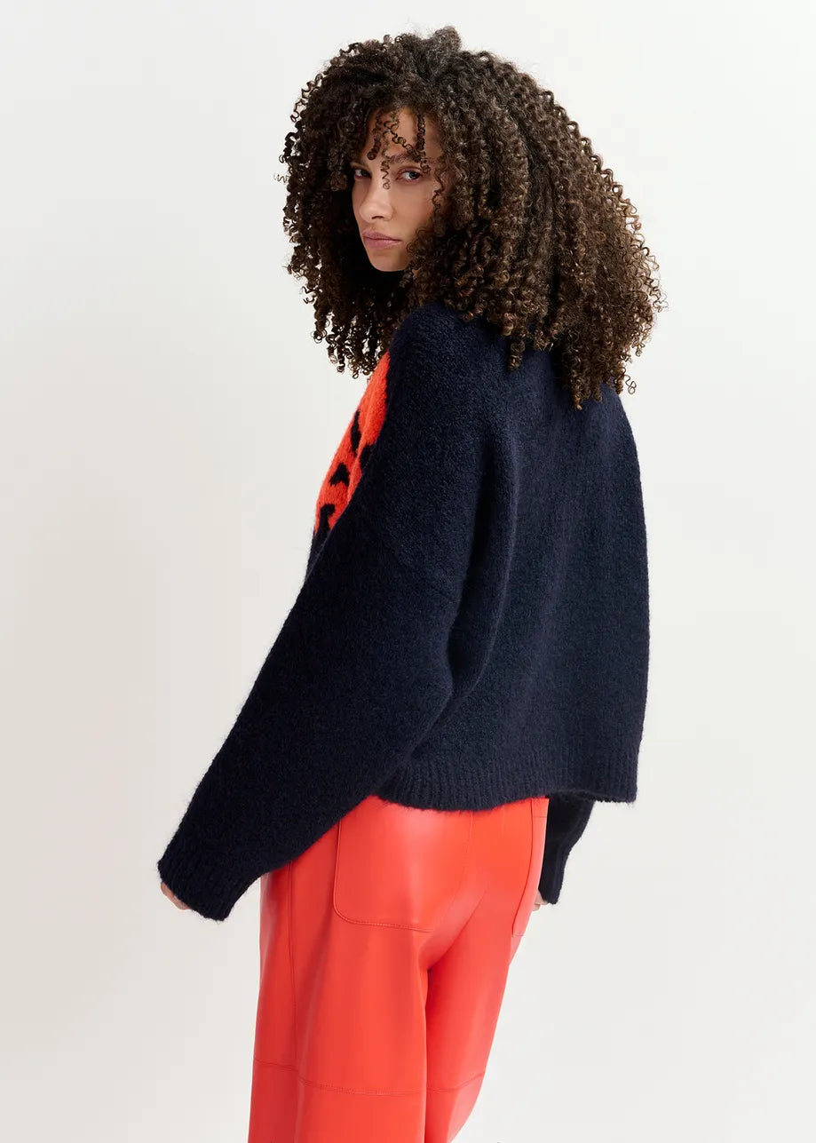 A person with curly hair wears an oversized navy and red Garcon Sweater by Essentiel Antwerp, paired with red pants. The person is standing against a plain background.