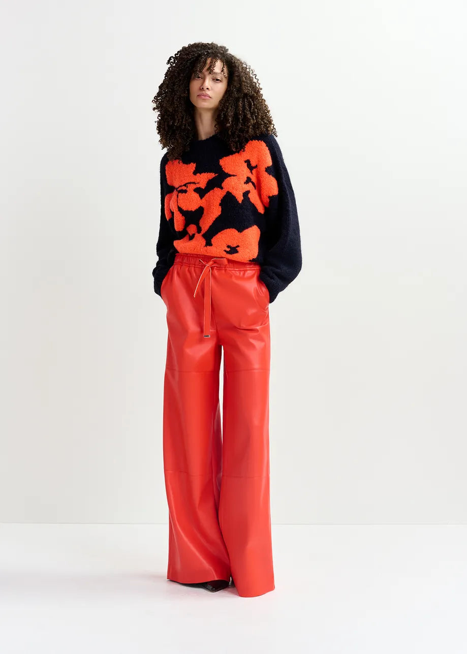 A person with curly hair stands against a plain background, wearing an oversized fit Garcon Sweater - Navy and Red by Essentiel Antwerp with large orange patterns and loose-fitting red pants, hands in pockets.