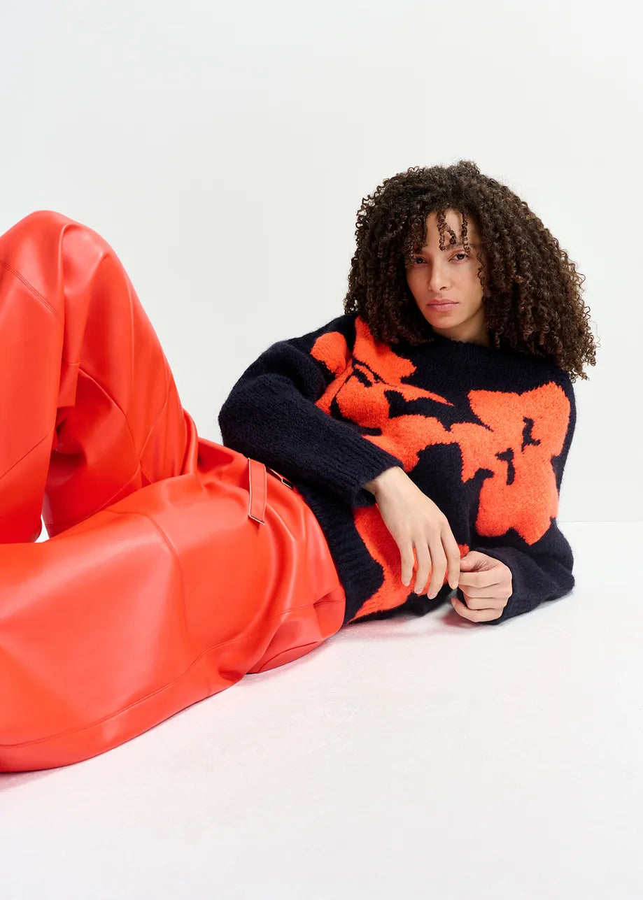 A person with curly hair in an oversized Garcon Sweater - Navy and Red by Essentiel Antwerp, paired with red leather pants, reclining on a white surface.