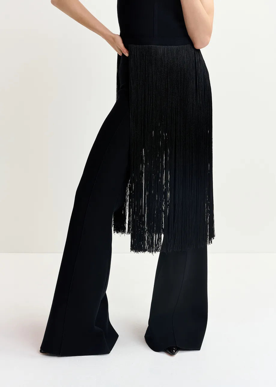 A person is wearing close-fitting Gattitude Knit Trousers in black by Essentiel Antwerp, featuring long black fringes on the sides that partially cover the pants. Only the lower torso and legs are visible.