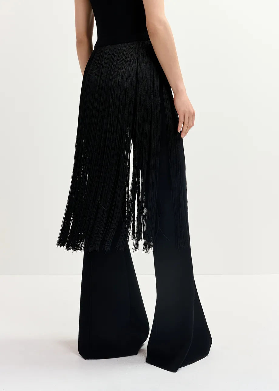 A person is wearing Essentiel Antwerp's Gattitude Knit Trousers in black, paired with a black fringe skirt overlay, set against a plain white background. The ensemble elegantly highlights the contrast between the loose silhouette of the trousers and the hint of close fit brought by the skirt overlay.