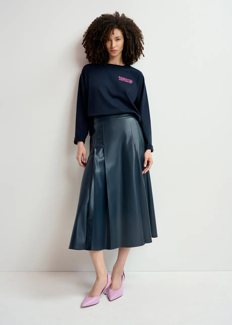 A woman stands against a plain background wearing a black long-sleeve top, the Geeuw Faux Leather Skirt in Navy by Essentiel Antwerp, and light pink shoes.