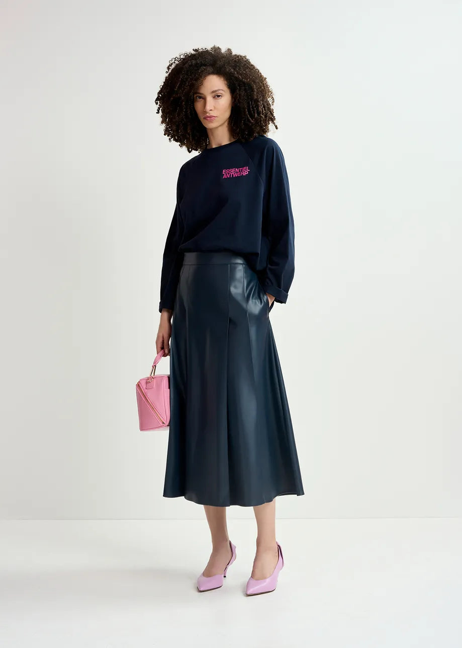 A person with curly hair is wearing a black long-sleeved shirt, a navy faux leather skirt from Essentiel Antwerp’s Geeuw collection, and pink shoes while holding a small pink handbag. They are standing against a plain white background.