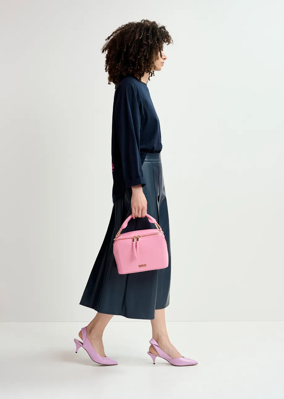 A person with curly hair walks in profile, wearing a black top, the Essentiel Antwerp Geeuw Faux Leather Skirt in navy, light pink heels, and carrying a light pink handbag. The background is a plain off-white wall.