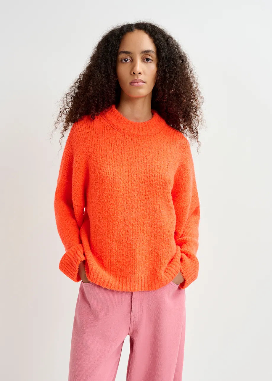 A person with long curly hair wears the Genlight Chunky Knit in Bright Orange by Essentiel Antwerp and pink pants, standing against a plain background.