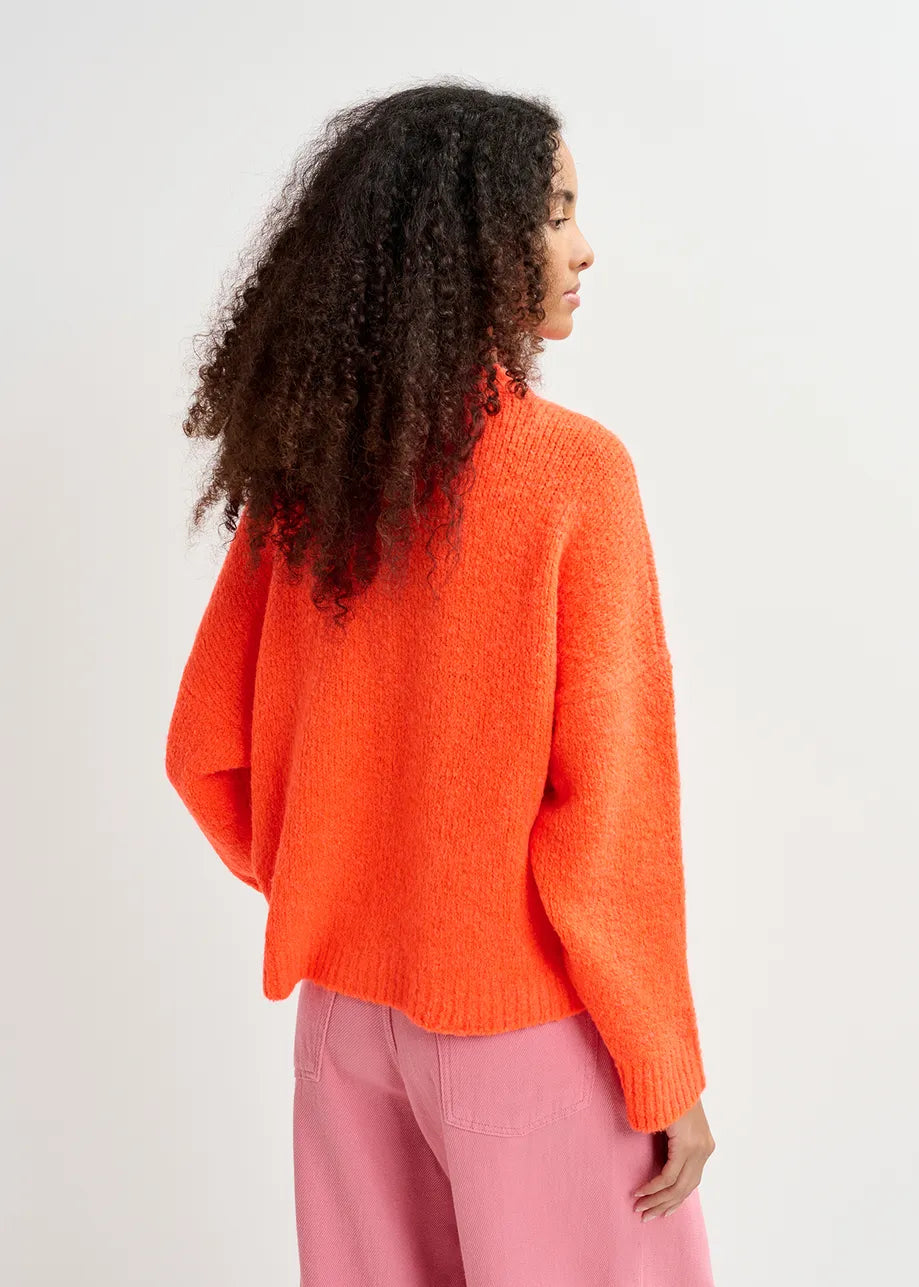 Person with curly hair wearing the Essentiel Antwerp Genlight Chunky Knit in bright orange and pink pants, standing against a white background, facing away from the camera.