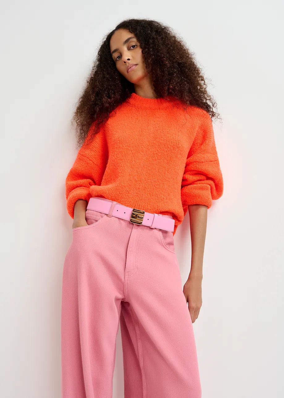 A person stands against a white background, wearing the Essentiel Antwerp Genlight Chunky Knit in Bright Orange and pink high-waisted pants with a matching pink belt.