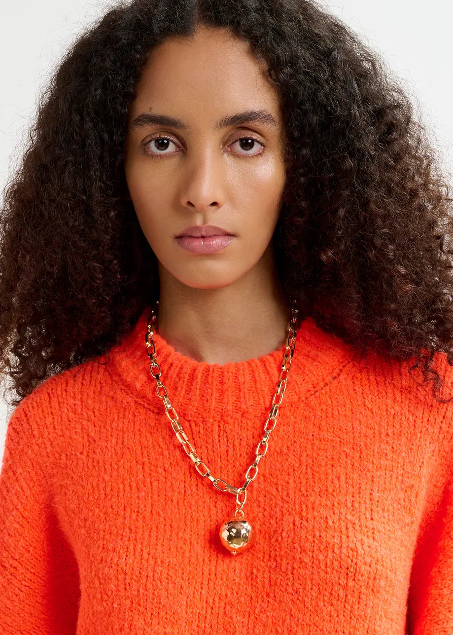 A person with curly hair wears the Essentiel Antwerp Genlight Chunky Knit in a bright orange hue and a gold chain necklace with a round pendant.