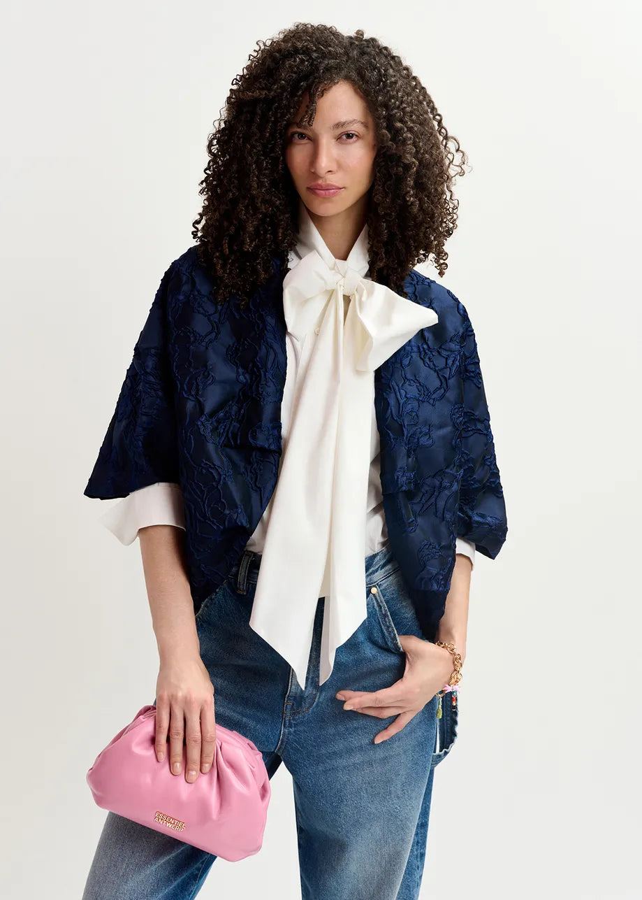 A person with curly hair is wearing a white blouse with a large bow, the Essentiel Antwerp Genuine Jacquard Jacket in navy, and blue jeans, while holding a pink clutch bag.