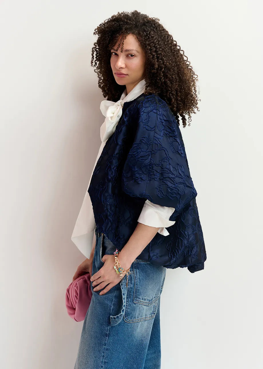 Person with curly hair wearing a Genuine Jacquard Jacket - Navy by Essentiel Antwerp, a white blouse with a large bow, and blue jeans, holding a pink clutch. Background is plain white.