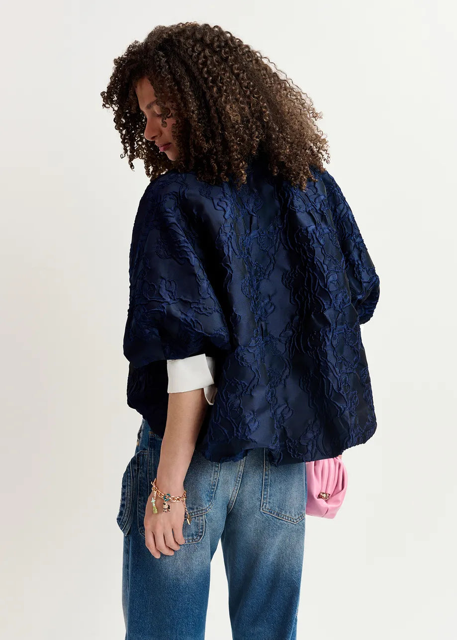 A person with curly hair is seen from behind, wearing an oversized jacket named the Genuine Jacquard Jacket in navy by Essentiel Antwerp, along with blue jeans and holding a pink purse.