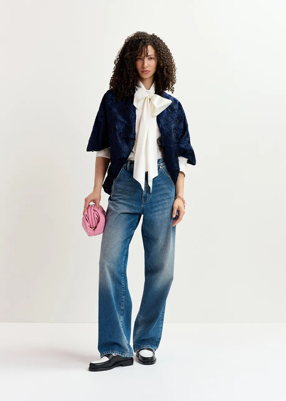 A person stands against a plain background wearing an oversized Genuine Jacquard Jacket in navy from Essentiel Antwerp, a white blouse with a large bow, wide-leg jeans, and black-and-white shoes, while holding a small pink clutch.