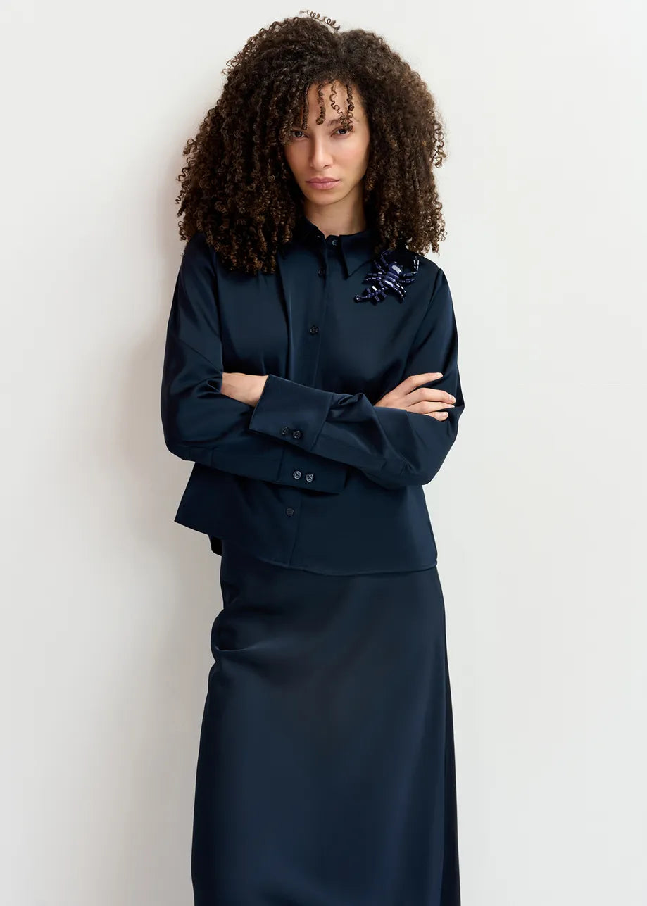 A woman with curly hair is standing against a white background, wearing the dark, long-sleeved Genvieve Satin Shirt - Navy from Essentiel Antwerp made from recycled polyester and a matching skirt. Her arms are crossed, and she is looking forward in relaxed fit attire.