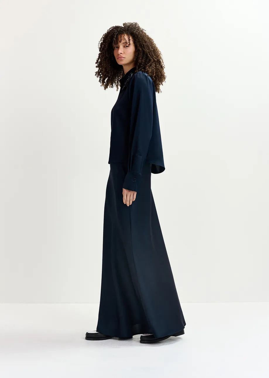 A person with curly hair stands against a plain background, wearing the Genvieve Satin Shirt in navy by Essentiel Antwerp. The long sleeves and high collar of the recycled polyester shirt create an air of effortless elegance, complemented by black shoes.