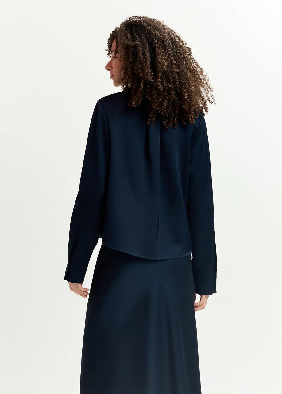 A person with curly hair is standing and facing away, wearing a relaxed fit navy blue long-sleeve Genvieve Satin Shirt from Essentiel Antwerp and a matching skirt made from recycled polyester.