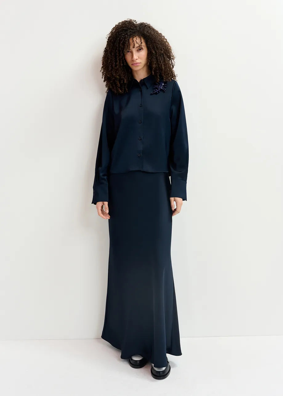 A woman with curly hair is wearing the Genvieve Satin Shirt in navy by Essentiel Antwerp, along with a matching long skirt made from recycled polyester, standing against a plain white background.