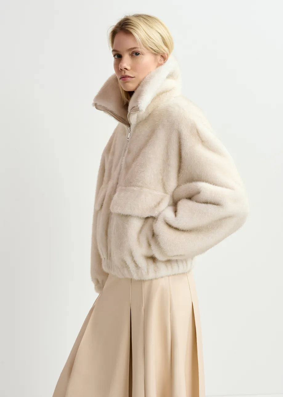 A person with blonde hair wears a white Genz Faux Fur Bomber Jacket by Essentiel Antwerp with an oversized bomber silhouette and a beige pleated skirt. Their hands are in the jacket pockets, and they look over their left shoulder.