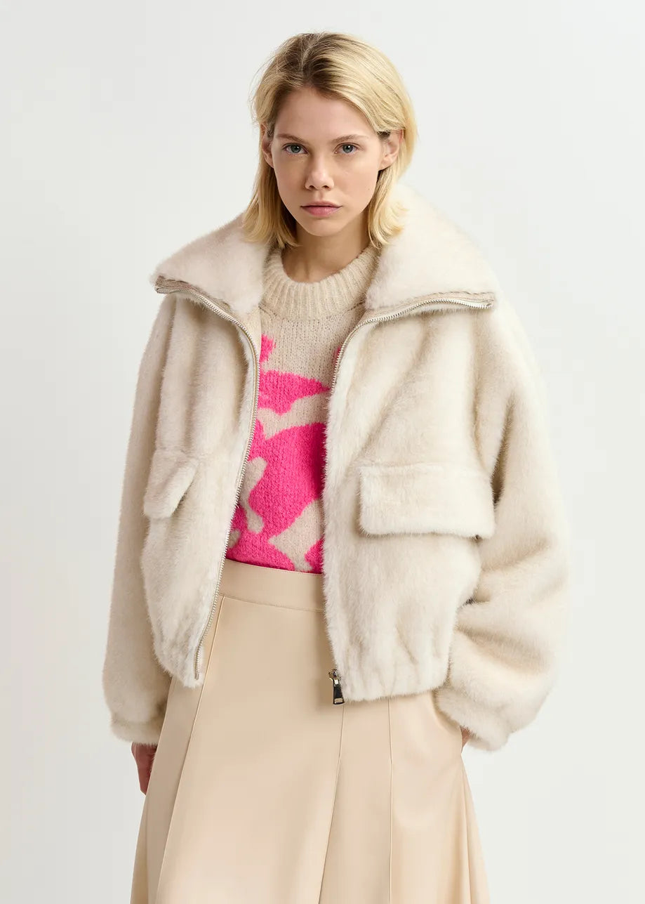 A person stands wearing a Genz Faux Fur Bomber Jacket in White by Essentiel Antwerp, with an oversized bomber silhouette, paired over a beige skirt and a beige sweater featuring a pink abstract pattern.