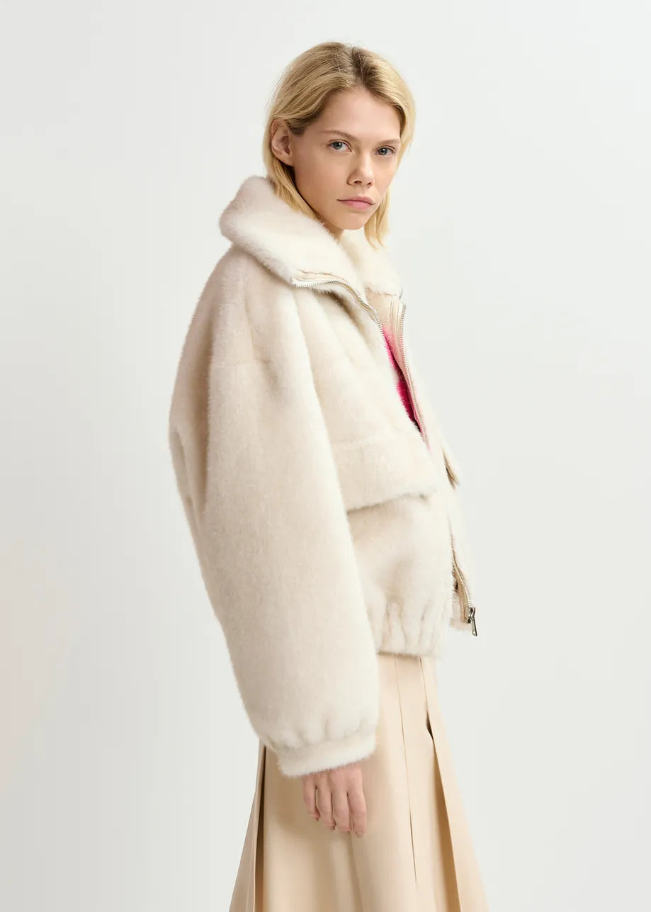 A person with blonde hair is wearing the Genz Faux Fur Bomber Jacket in white from Essentiel Antwerp and a light beige skirt, standing against a plain light background, with the oversized bomber silhouette adding to the relaxed vibe.